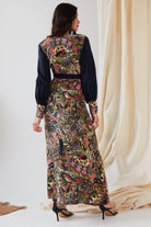 A woman faces away, wearing the Sarvin Hand Painted Long Sleeve Maxi Dress featuring a colorful paisley and floral design with dark sleeves and waistband, paired with black heels before softly draped light fabric.