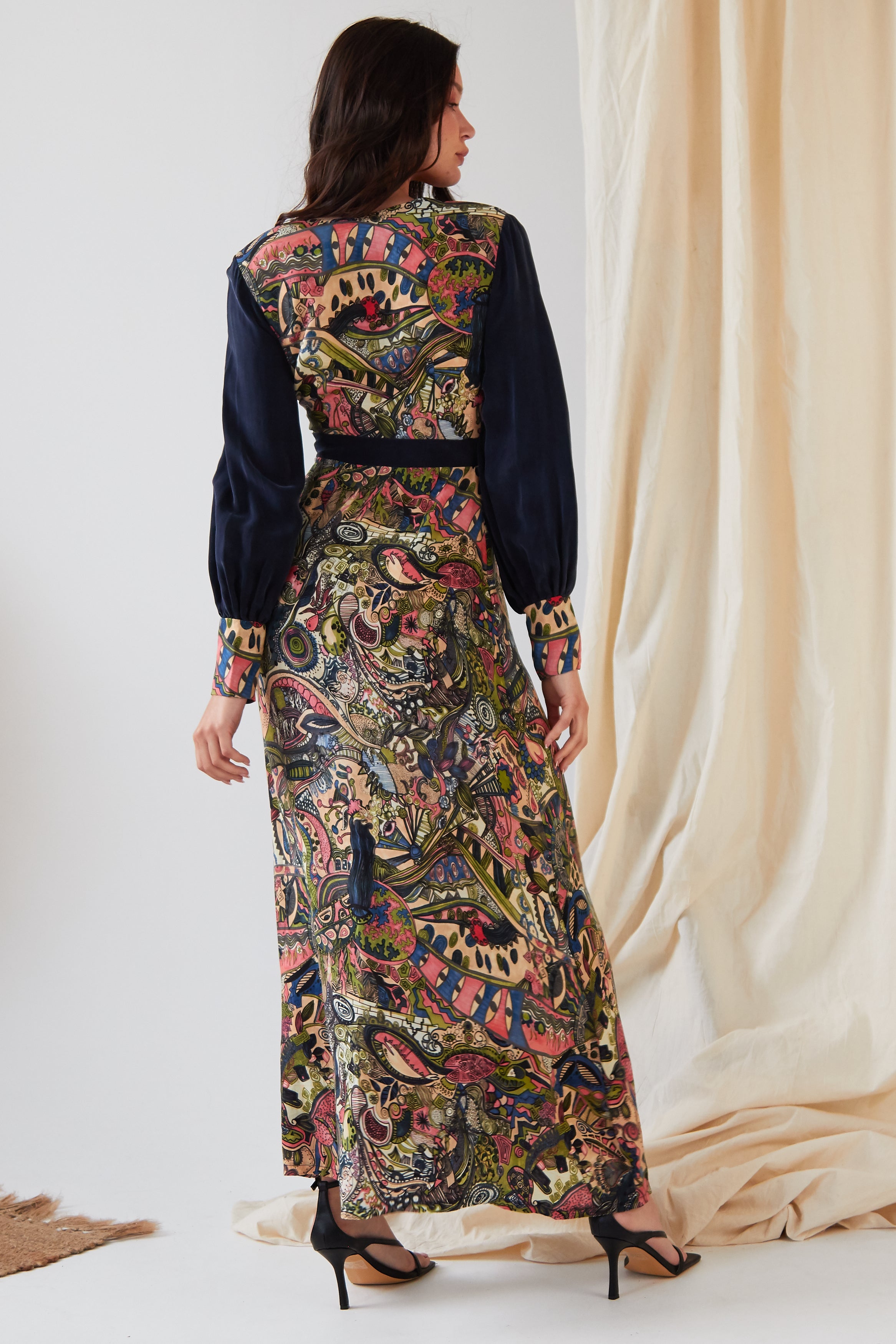 A woman faces away, wearing the Sarvin Hand Painted Long Sleeve Maxi Dress featuring a colorful paisley and floral design with dark sleeves and waistband, paired with black heels before softly draped light fabric.