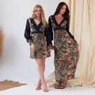 Two women pose indoors in Sarvin's Hand Painted Long Sleeve Mini Dress and a Persian print mini dress—both with navy tops and vibrant patterned skirts. Beige drapes and pampas grass create a soft, elegant backdrop.