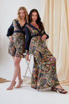 Two women pose together in paisley-print dresses from Sarvin, featuring the Hand painted Long Sleeve Maxi Dress, set against a neutral draped fabric background.