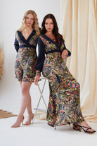 Two women pose indoors: one stands in the Sarvin Hand Painted Long Sleeve Mini Dress, while the other sits on a high stool wearing a matching long dress. Both feature dark fitted tops and vibrant, hand-painted skirts.