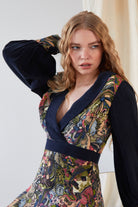 A woman with long, wavy blonde hair wears the Sarvin Hand Painted Long Sleeve Mini Dress featuring colorful, intricate patterns. She stands in front of a light background, looking to the side with one hand behind her head.