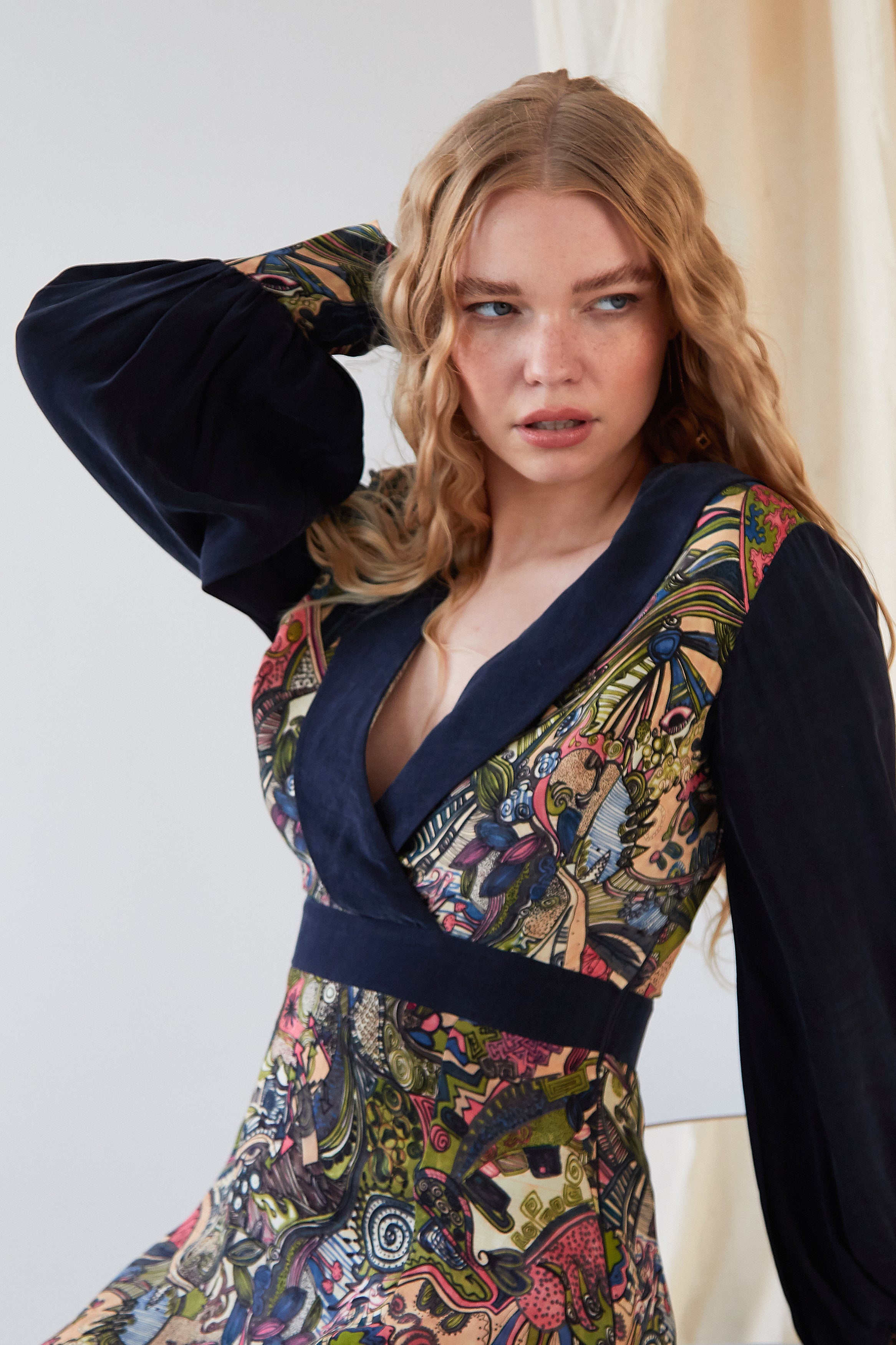 A woman with long, wavy blonde hair wears the Sarvin Hand Painted Long Sleeve Mini Dress featuring colorful, intricate patterns. She stands in front of a light background, looking to the side with one hand behind her head.