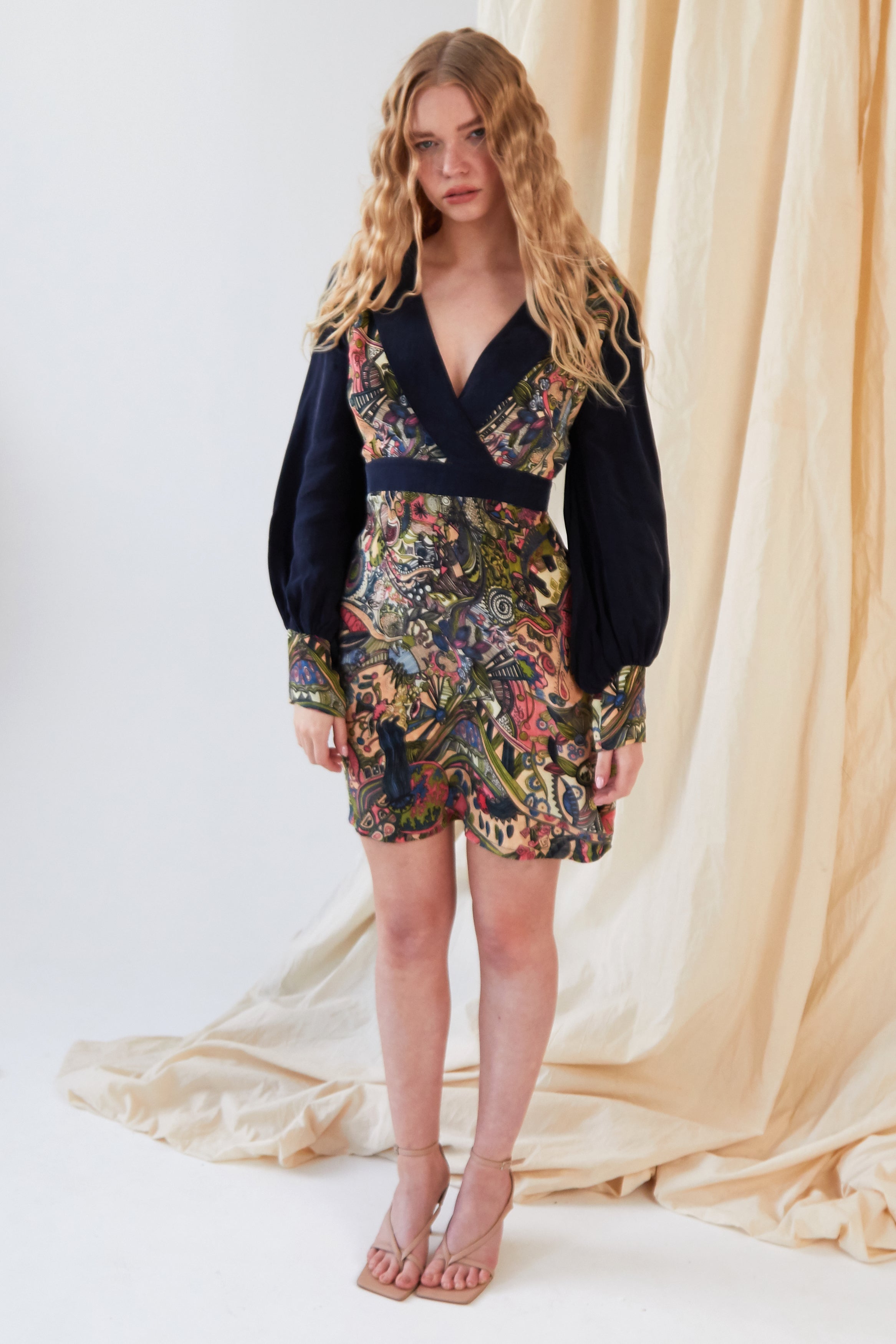 A woman with long wavy blonde hair stands indoors on a white floor before beige drapes, wearing the Sarvin Hand Painted Long Sleeve Mini Dress and beige strappy heels.