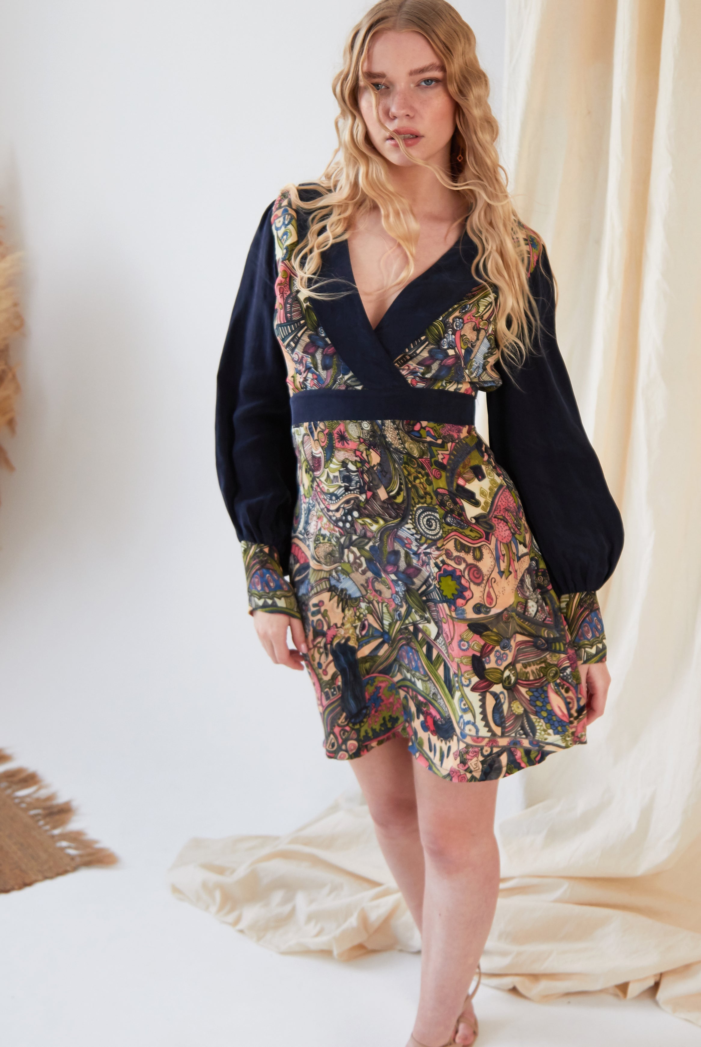 A woman with long, wavy blonde hair wears the Sarvin Hand Painted Long Sleeve Mini Dress, showcasing black sleeves and a vibrant, intricate Persian-inspired print. She stands indoors near cream-colored fabric and a woven rug.