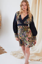 A woman with long, wavy blonde hair wears the Sarvin Hand Painted Long Sleeve Mini Dress, showcasing black sleeves and a vibrant, intricate Persian-inspired print. She stands indoors near cream-colored fabric and a woven rug.