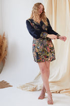 A woman with long wavy blonde hair wears the Sarvin Hand painted Long Sleeve Mini Dress, featuring a colorful patterned skirt, standing on a light floor with flowing fabric and pampas grass in the background.
