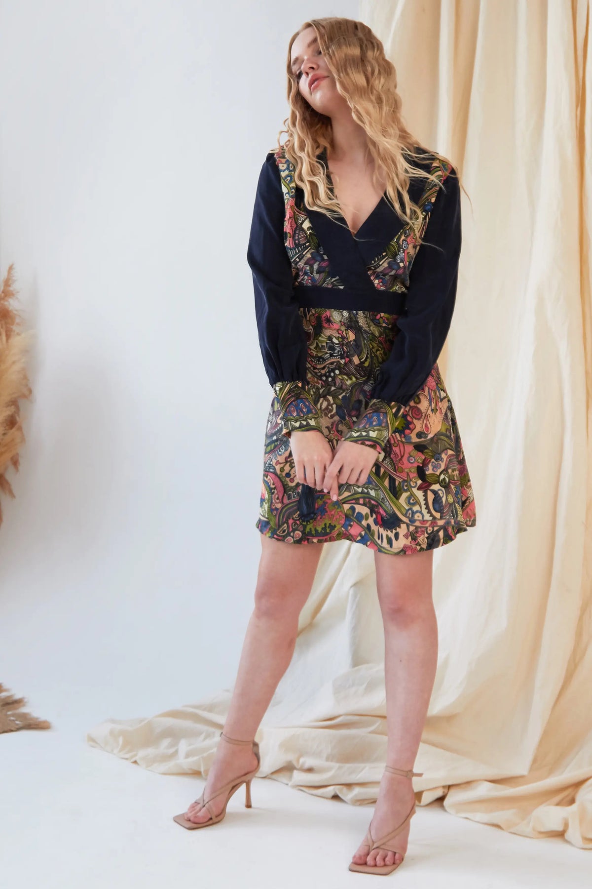 A woman with long, wavy blonde hair smiles indoors, wearing the Sarvin Hand painted Long Sleeve Mini Dress featuring a dark top and a colorful patterned skirt. She stands with her hands held loosely in front, gazing slightly to the side.