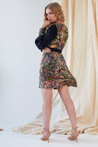 A woman stands sideways before cream drapes, wearing the Sarvin Hand Painted Long Sleeve Mini Dress with dark sleeves and nude heels. She looks over her shoulder, one hand near her face.