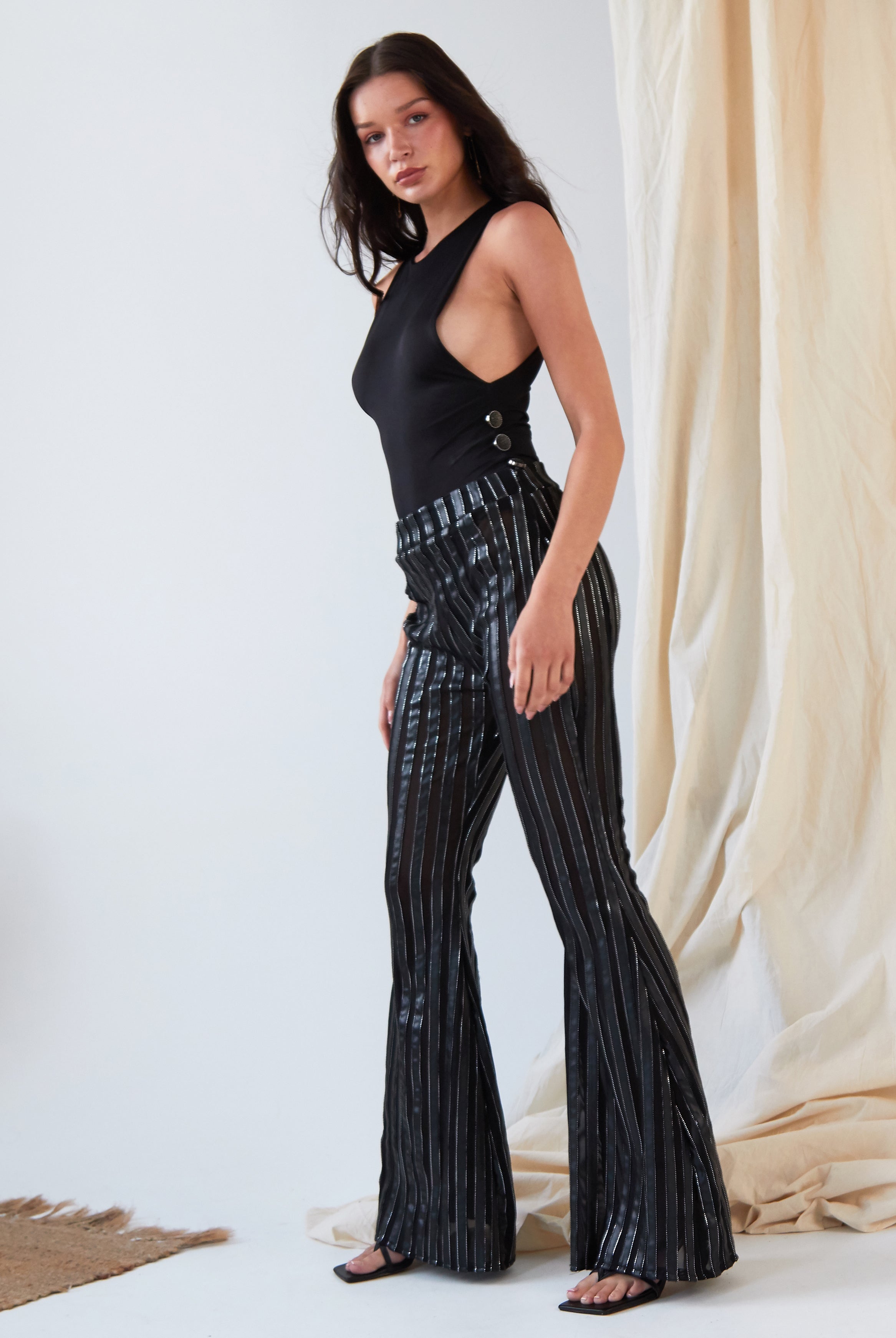 A woman indoors wears the Sarvin Dropped Armhole Bodysuit with high-waisted, black flared pants featuring shiny vertical stripes. She stands sideways, looking at the camera.