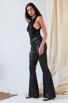 A woman indoors wears the Sarvin Dropped Armhole Bodysuit with high-waisted, black flared pants featuring shiny vertical stripes. She stands sideways, looking at the camera.