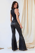 A woman faces slightly away from the camera wearing the Sarvin Dropped Armhole Bodysuit, a sleeveless black party piece, paired with shiny black flared pants with vertical stripes. Beige curtains serve as the backdrop.