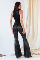 A woman stands facing away in front of light draped fabric, wearing Sarvin's Dropped Armhole Bodysuit and shiny black flare pants with vertical silver stripes.