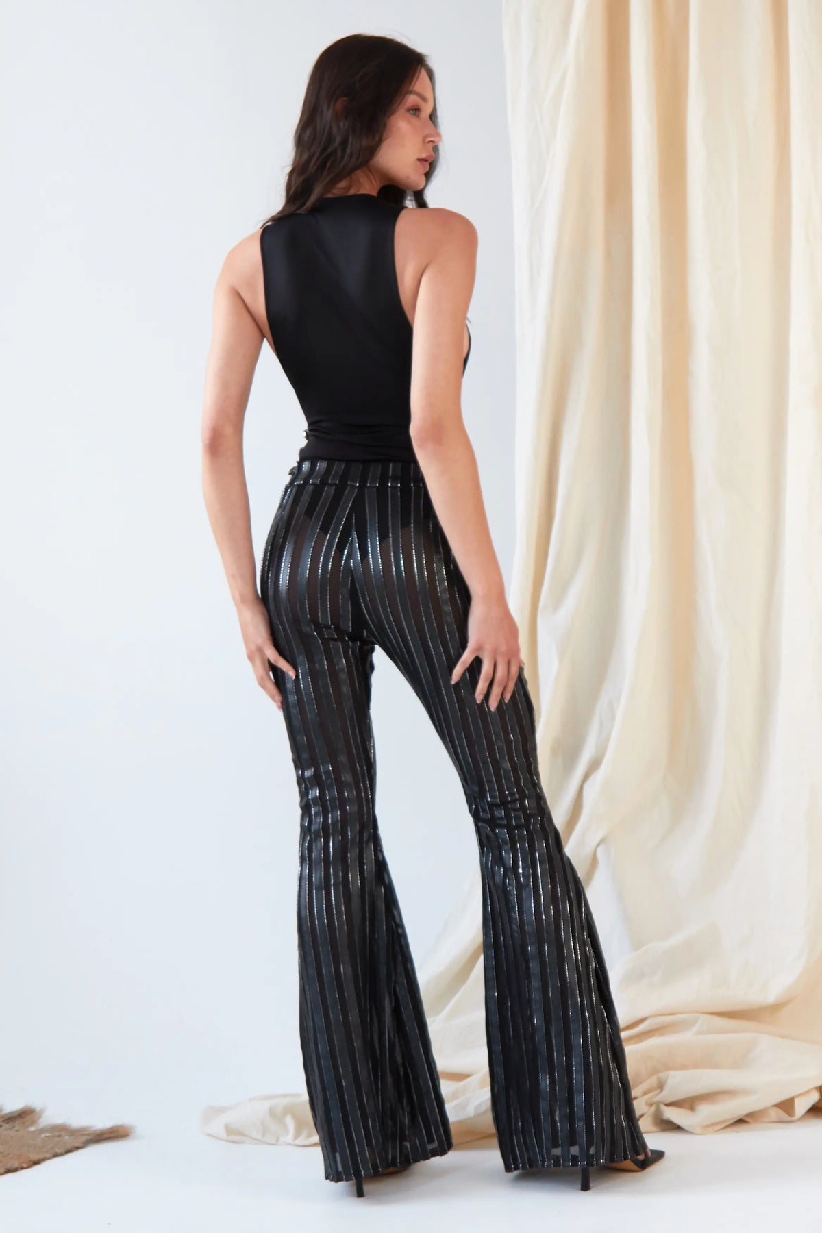 A woman stands facing away in front of light draped fabric, wearing Sarvin's Dropped Armhole Bodysuit and shiny black flare pants with vertical silver stripes.