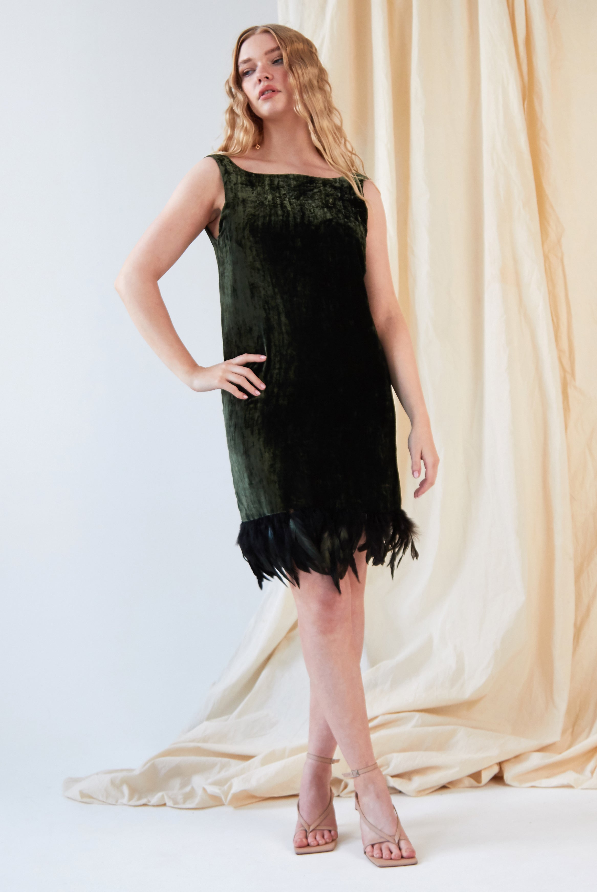 A woman poses before cream drapes in Sarvin's Silk Velvet Feather Trim Dress, a sleeveless forest green cocktail dress with an ostrich feather hem, paired with nude strappy heels and one hand on her hip, looking slightly to the side.