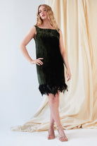 A woman poses before cream drapes in Sarvin's Silk Velvet Feather Trim Dress, a sleeveless forest green cocktail dress with an ostrich feather hem, paired with nude strappy heels and one hand on her hip, looking slightly to the side.