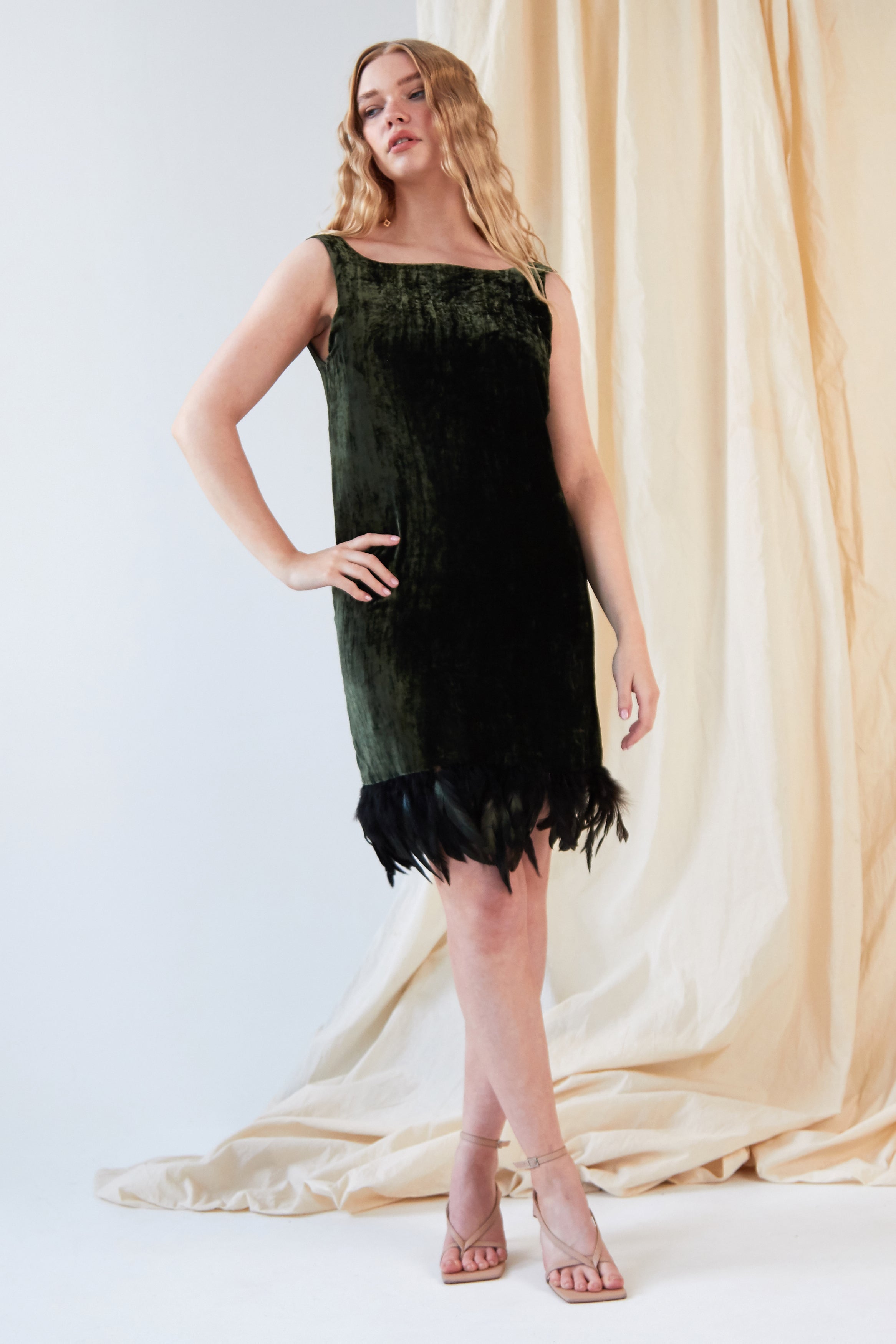 A woman poses before cream drapes in Sarvin's Silk Velvet Feather Trim Dress, a sleeveless forest green cocktail dress with an ostrich feather hem, paired with nude strappy heels and one hand on her hip, looking slightly to the side.