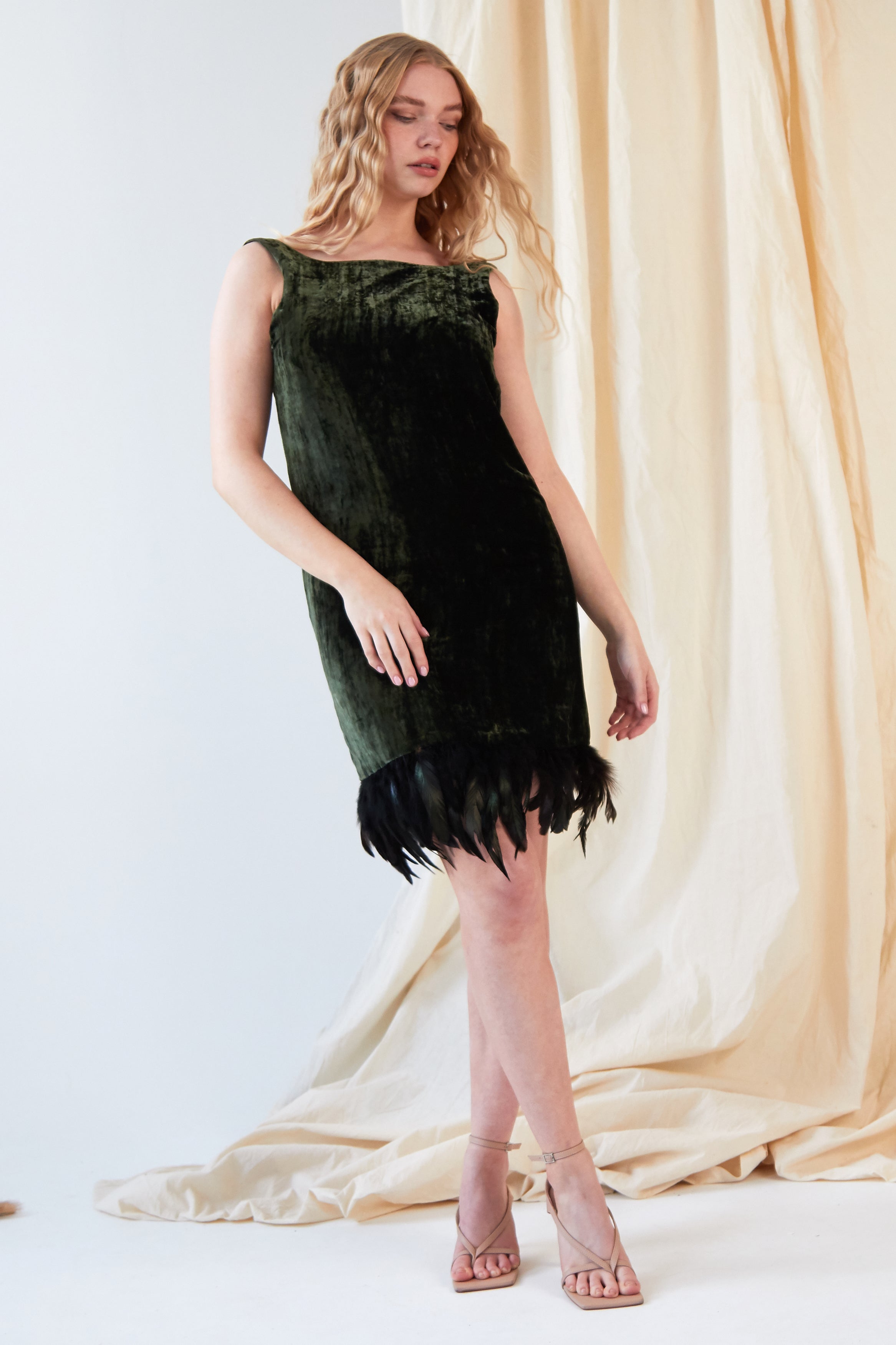 A woman poses in front of beige drapes, wearing the Sarvin Silk Velvet Feather Trim Dress in forest green with an ostrich feather hem and nude strappy heels. Her long wavy blonde hair frames her face as she looks ahead neutrally.