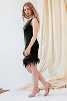A woman with long wavy hair wears the Sarvin Silk Velvet Feather Trim Dress, a sleeveless forest green cocktail dress with an ostrich feather hem, and nude heels, standing sideways before a draped beige fabric backdrop.