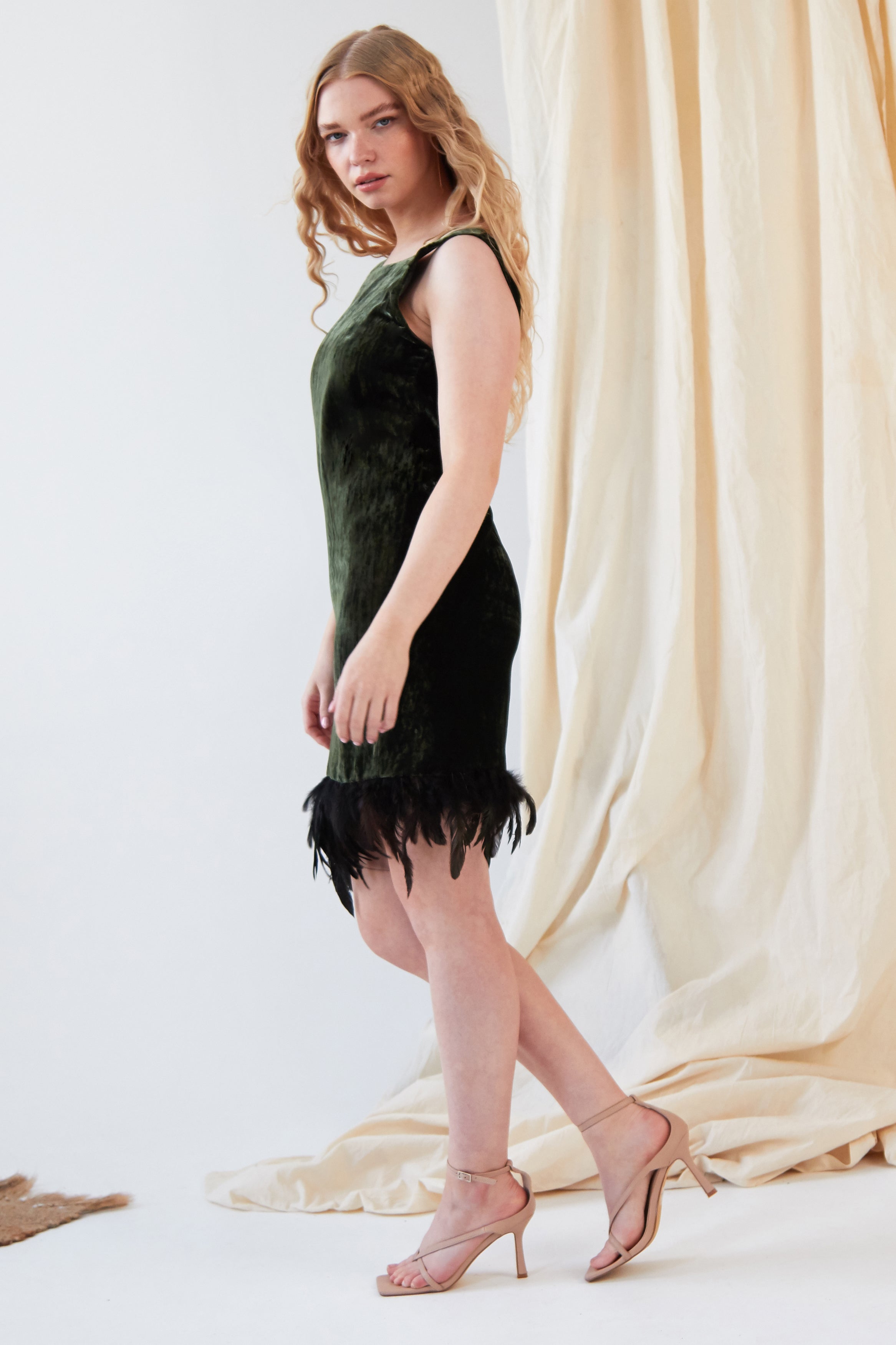 A woman with long wavy hair wears the Sarvin Silk Velvet Feather Trim Dress, a sleeveless forest green cocktail dress with an ostrich feather hem, and nude heels, standing sideways before a draped beige fabric backdrop.