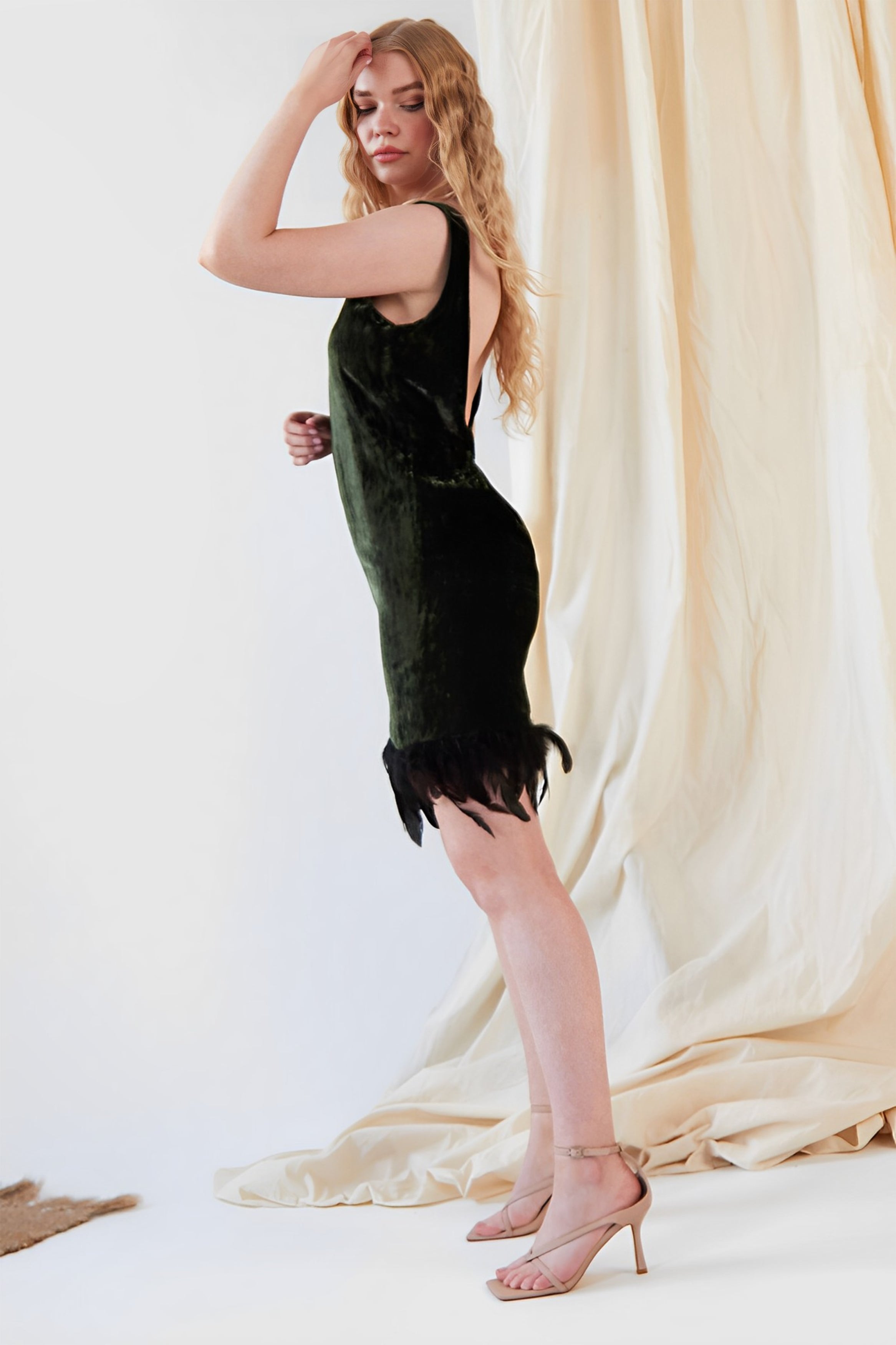 A woman with long wavy blonde hair wears the Sarvin Silk Velvet Feather Trim Dress and nude high-heeled sandals, standing in profile against a cream-colored draped fabric backdrop.
