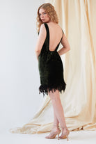 A person stands in profile wearing the Sarvin Silk Velvet Feather Trim Dress featuring a low back and ostrich feather hem, paired with nude heeled sandals. A cream-colored fabric is draped in the background.