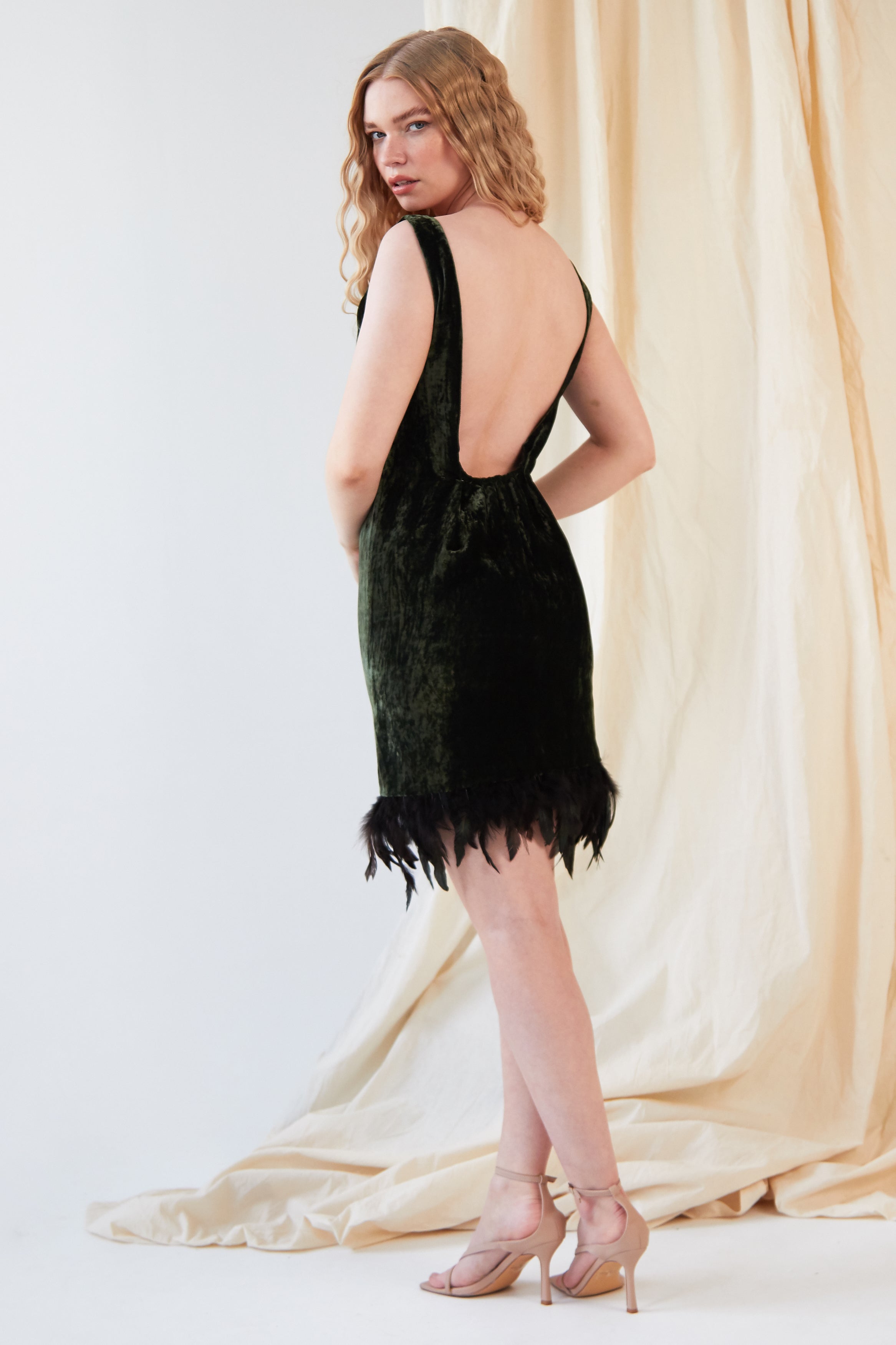 A person stands in profile wearing the Sarvin Silk Velvet Feather Trim Dress featuring a low back and ostrich feather hem, paired with nude heeled sandals. A cream-colored fabric is draped in the background.