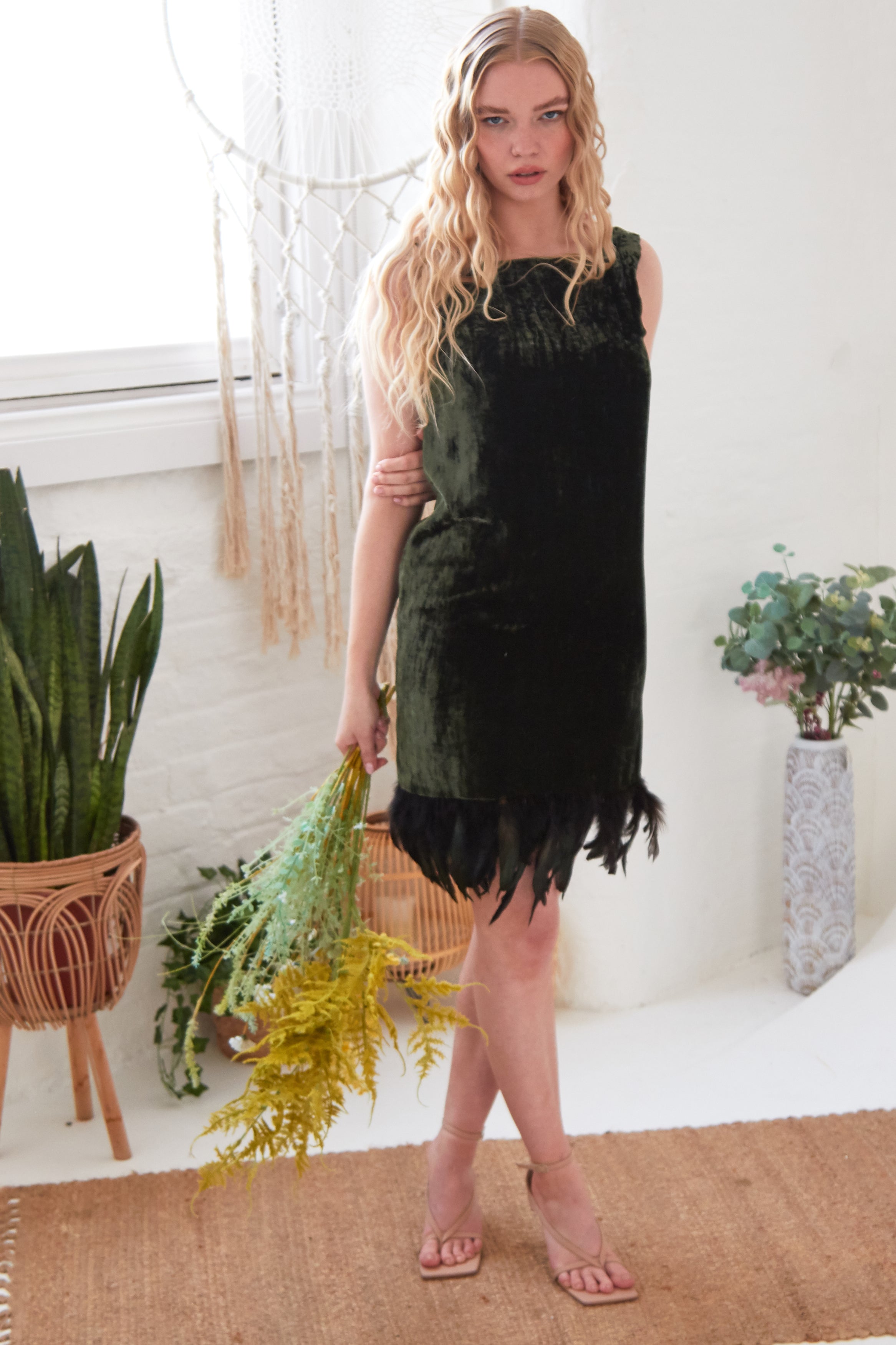 A woman with long blonde hair stands indoors wearing the Sarvin Silk Velvet Feather Trim Dress. She holds a bouquet of yellow-green foliage, standing on a woven rug near potted plants and a white brick wall.