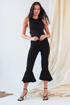 A woman poses indoors in the Sarvin Dropped Armhole Bodysuit, featuring flared legs and paired with black strappy heels. She stands against beige draped fabric—a chic, effortless look perfect for any party.