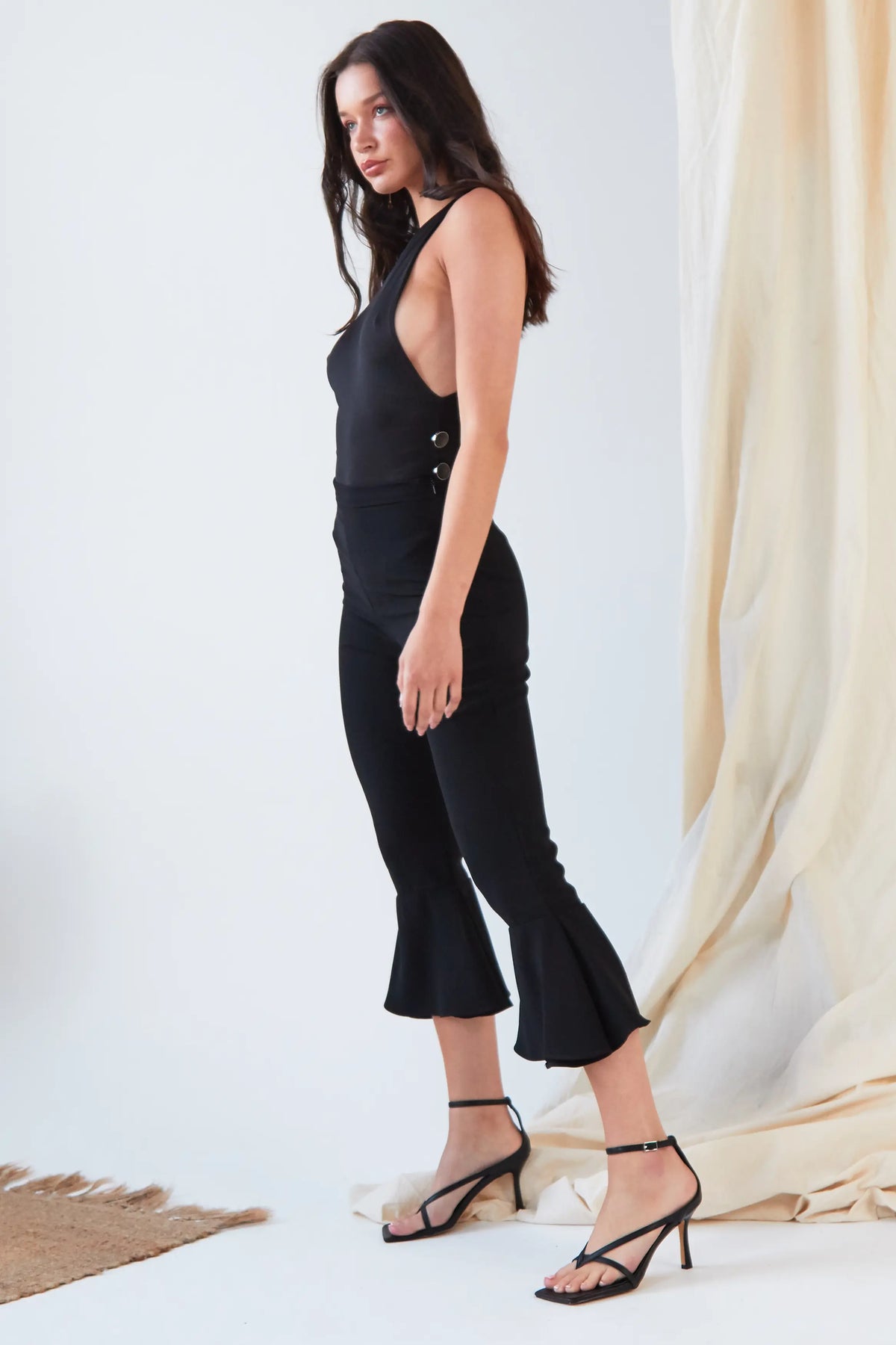 A woman stands indoors against a light background, wearing the Sarvin Dropped Armhole Bodysuit styled with fitted flared pants and black heeled sandals. Her hair is loose, and she gazes softly to the side.