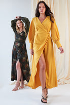 Two women model wrap dresses. The woman in front wears the Sarvin Mustard Wrap Maxi Dress with a deep V-neck and high slit, while the woman behind stuns in a dark, vintage-style satin dress with bishop sleeves and a matching slit.