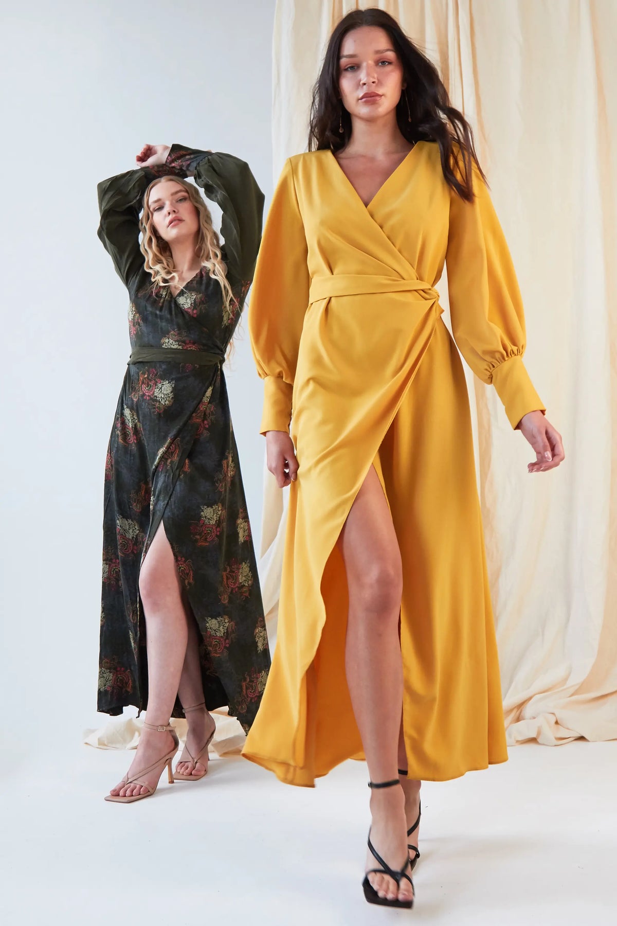 Two women model wrap dresses. The woman in front wears the Sarvin Mustard Wrap Maxi Dress with a deep V-neck and high slit, while the woman behind stuns in a dark, vintage-style satin dress with bishop sleeves and a matching slit.