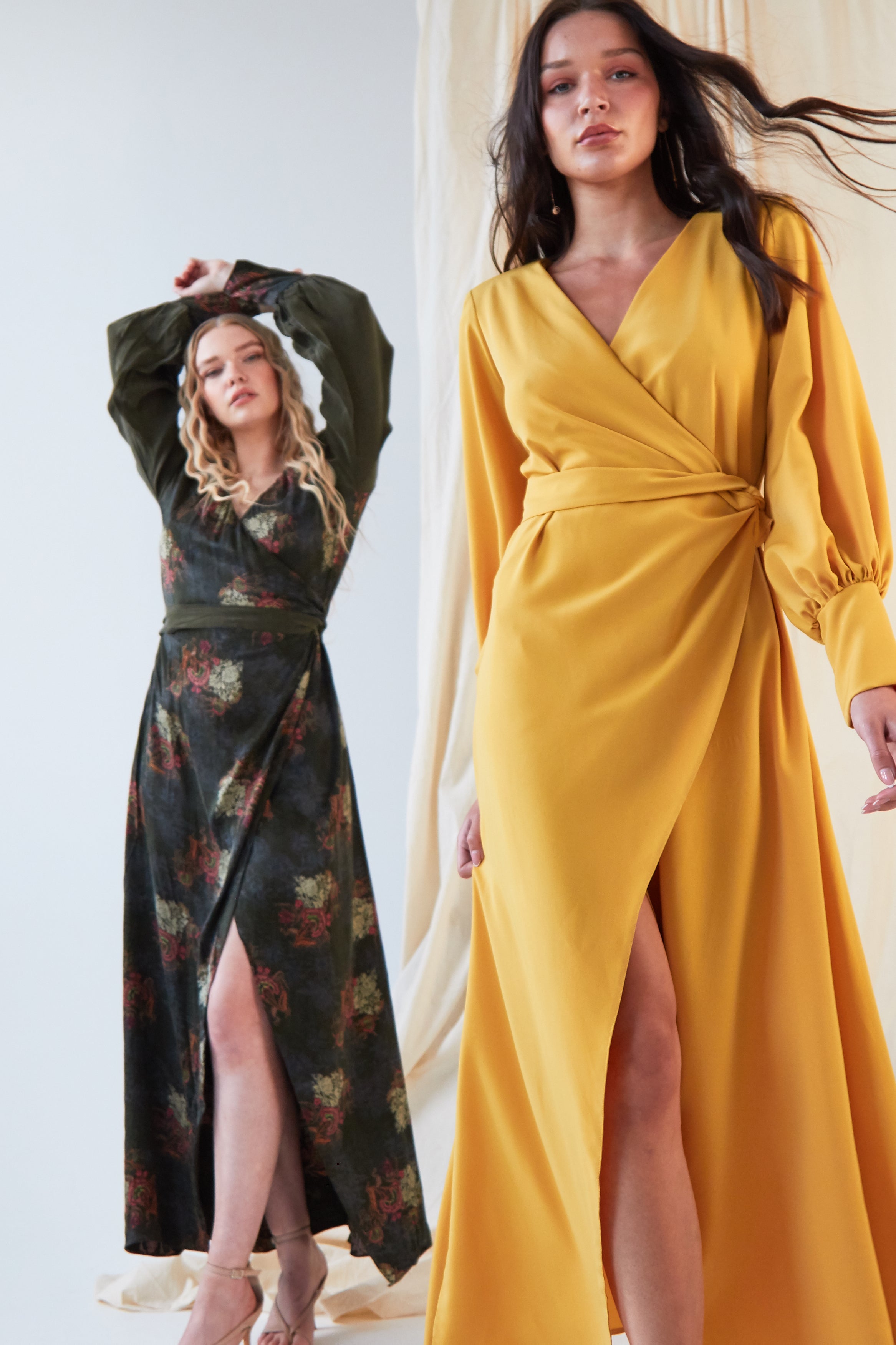 Two women stand indoors in Sarvin dresses. The woman in front wears the Mustard Wrap Maxi Dress with long sleeves, while the other wears a dark floral satin dress with olive green bishop sleeves. Both styles feature front slits.