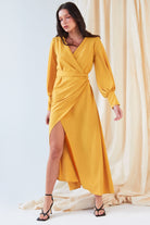 A woman poses before cream drapes in the Sarvin Mustard Wrap Maxi Dress, featuring vintage-inspired satin, balloon sleeves, a deep V-neck, and an asymmetrical hem, styled with black strappy high-heeled sandals.