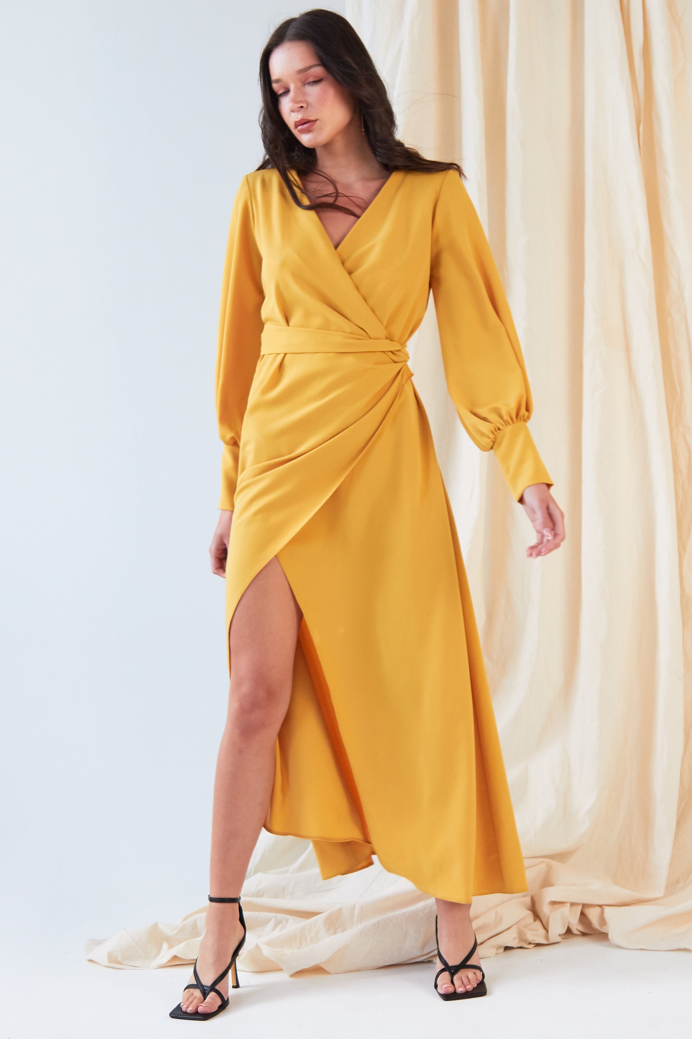 A woman poses before cream drapes in the Sarvin Mustard Wrap Maxi Dress, featuring vintage-inspired satin, balloon sleeves, a deep V-neck, and an asymmetrical hem, styled with black strappy high-heeled sandals.