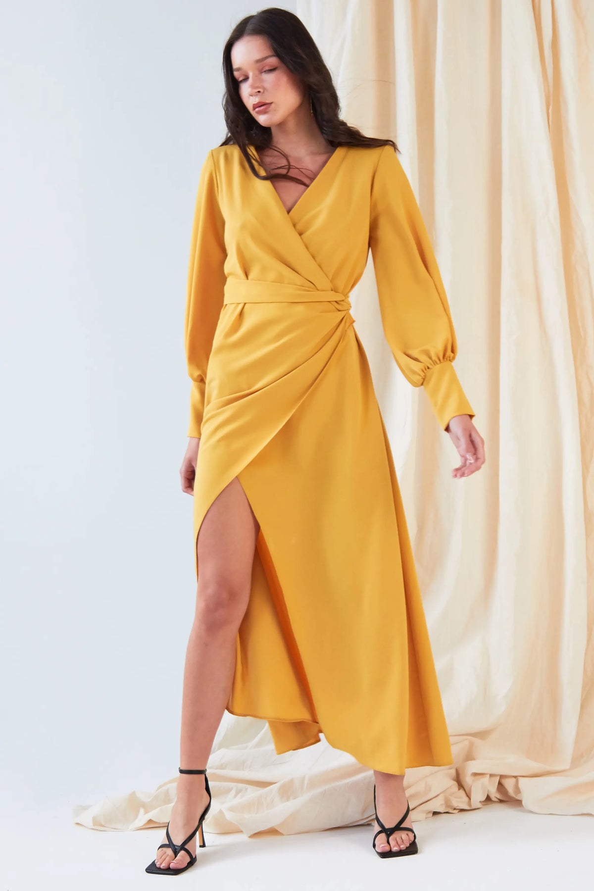 A woman poses before cream drapes in the Sarvin Mustard Wrap Maxi Dress, featuring vintage-inspired satin, balloon sleeves, a deep V-neck, and an asymmetrical hem, styled with black strappy high-heeled sandals.