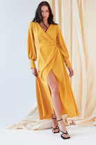 A woman models the Sarvin Mustard Wrap Maxi Dress, a vintage-inspired satin maxi with a front slit, styled with black strappy heels. She poses before a neutral draped backdrop, her long hair worn loose.