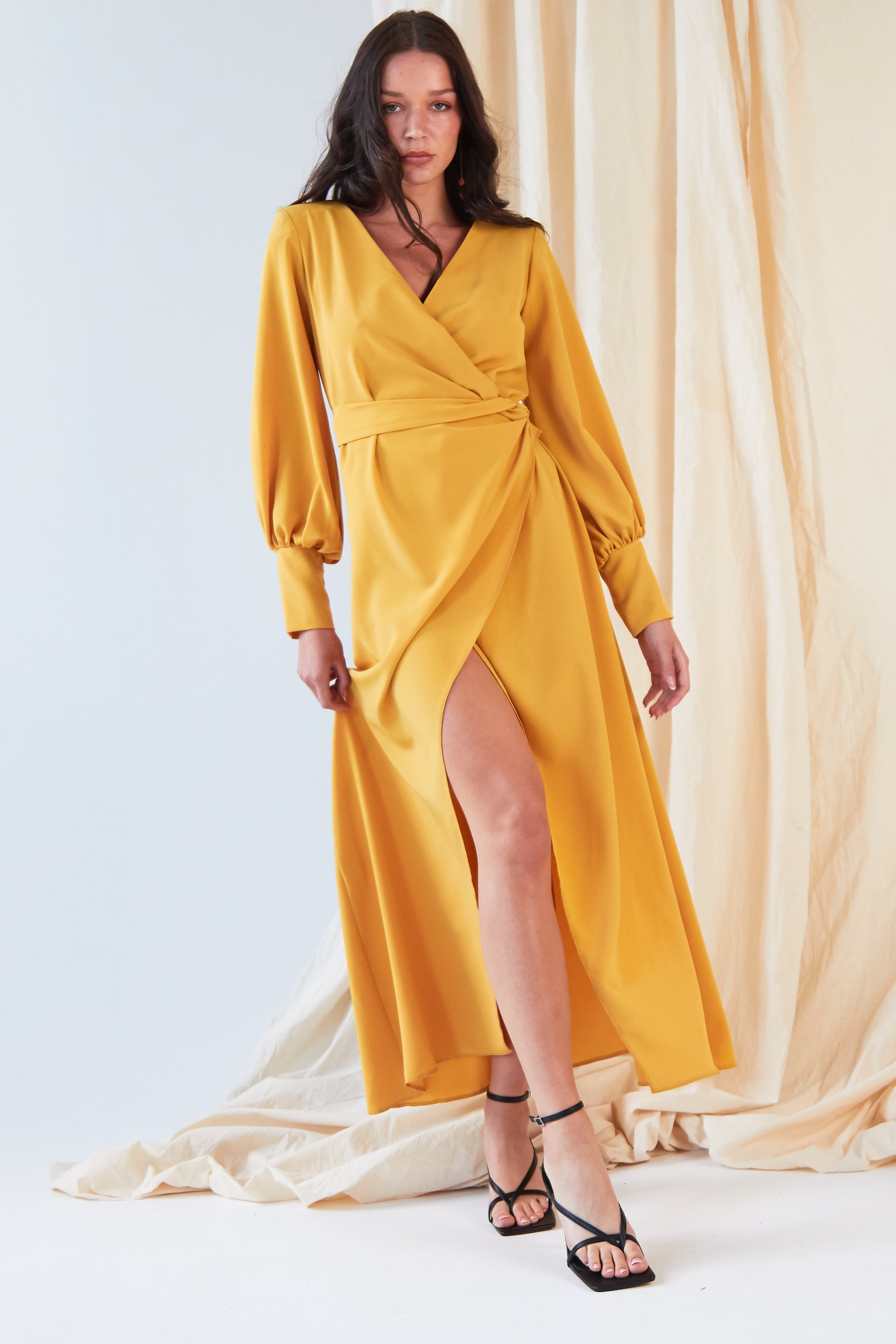 A woman models the Sarvin Mustard Wrap Maxi Dress, a vintage-inspired satin maxi with a front slit, styled with black strappy heels. She poses before a neutral draped backdrop, her long hair worn loose.