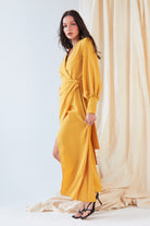 A woman models the Sarvin Mustard Wrap Maxi Dress, featuring balloon bishop sleeves and a thigh-high slit, in front of beige draped fabric. She pairs this vintage-inspired look with black strappy heels.