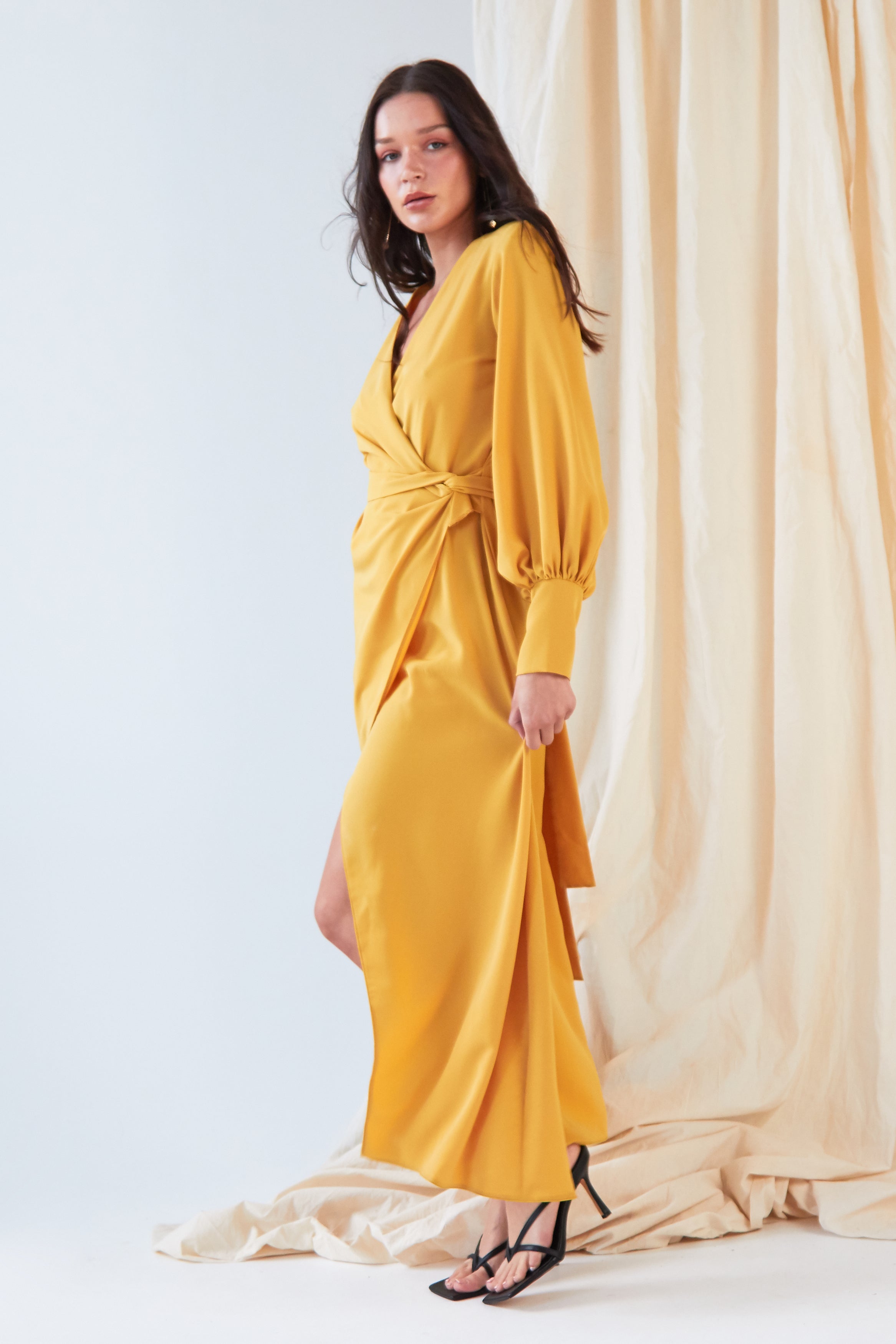 A woman models the Sarvin Mustard Wrap Maxi Dress, featuring balloon bishop sleeves and a thigh-high slit, in front of beige draped fabric. She pairs this vintage-inspired look with black strappy heels.