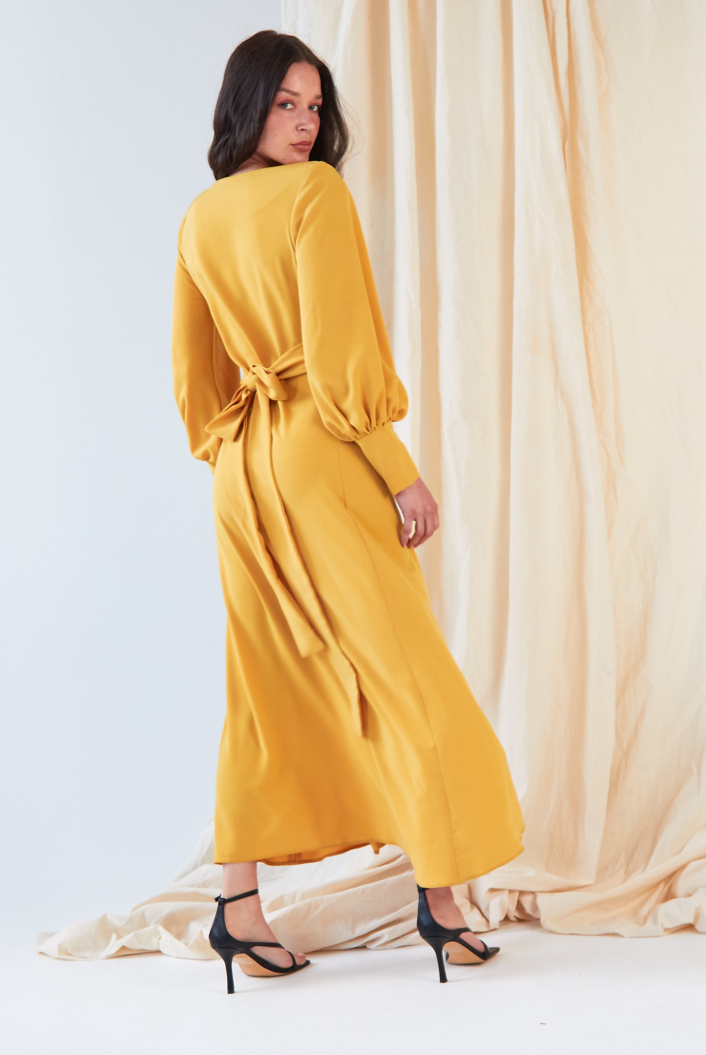 A woman wearing the Sarvin Mustard Wrap Maxi Dress with vintage-inspired balloon bishop sleeves stands before a cream curtain, looking over her shoulder and pairing the dress with black high-heeled sandals.