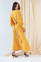 A woman wearing the Sarvin Mustard Wrap Maxi Dress with vintage-inspired balloon bishop sleeves stands before a cream curtain, looking over her shoulder and pairing the dress with black high-heeled sandals.
