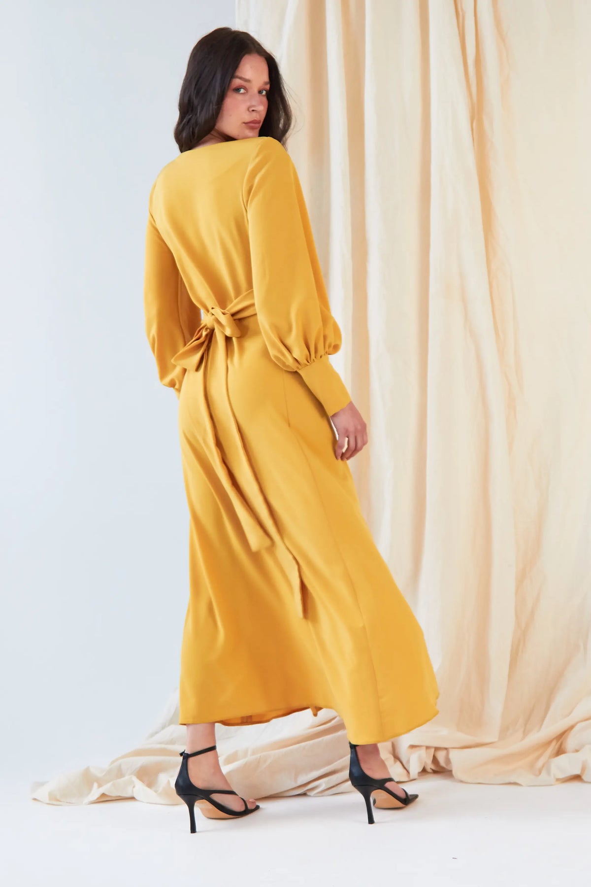 A woman wearing the Sarvin Mustard Wrap Maxi Dress with vintage-inspired balloon bishop sleeves stands before a cream curtain, looking over her shoulder and pairing the dress with black high-heeled sandals.