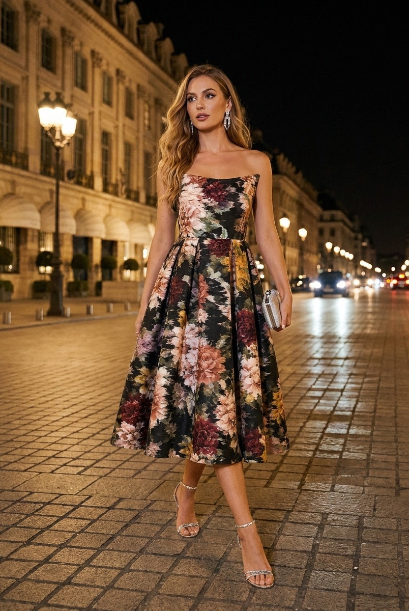 A woman in the Sarvin Golshifteh Floral Corset Midi Dress and heels walks on a cobblestone street at night, holding a small clutch, her pleated skirt swaying as she passes historic buildings lit by streetlights.