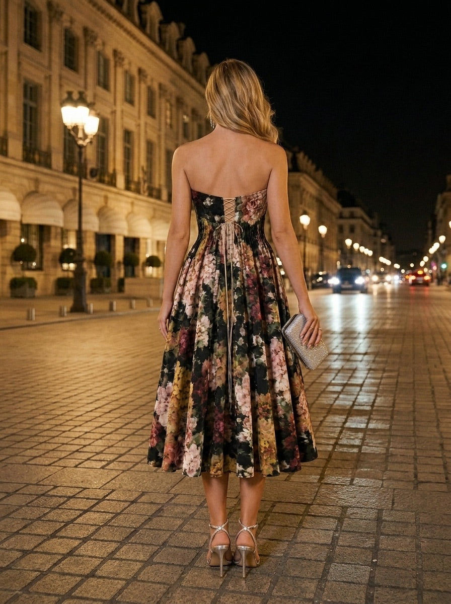 A woman with long blonde hair stands on a cobblestone street at night, illuminated by streetlights. She wears the Golshifteh Floral Corset Midi Dress by Sarvin and silver high heels, holding a clutch while facing away from the camera.