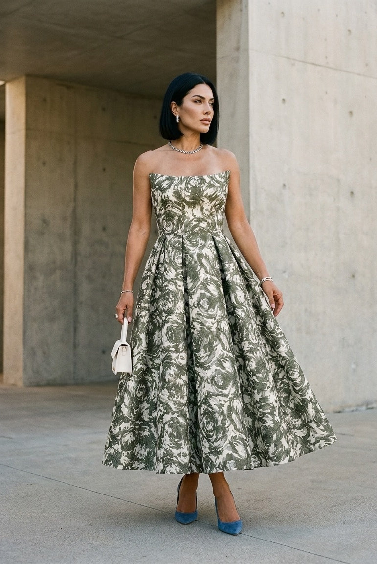 A woman stands outdoors against a concrete wall, wearing the Sarvin Tala Corset Midi Dress, styled with straight black hair, jewelry, blue high heels, and holding a small white purse.