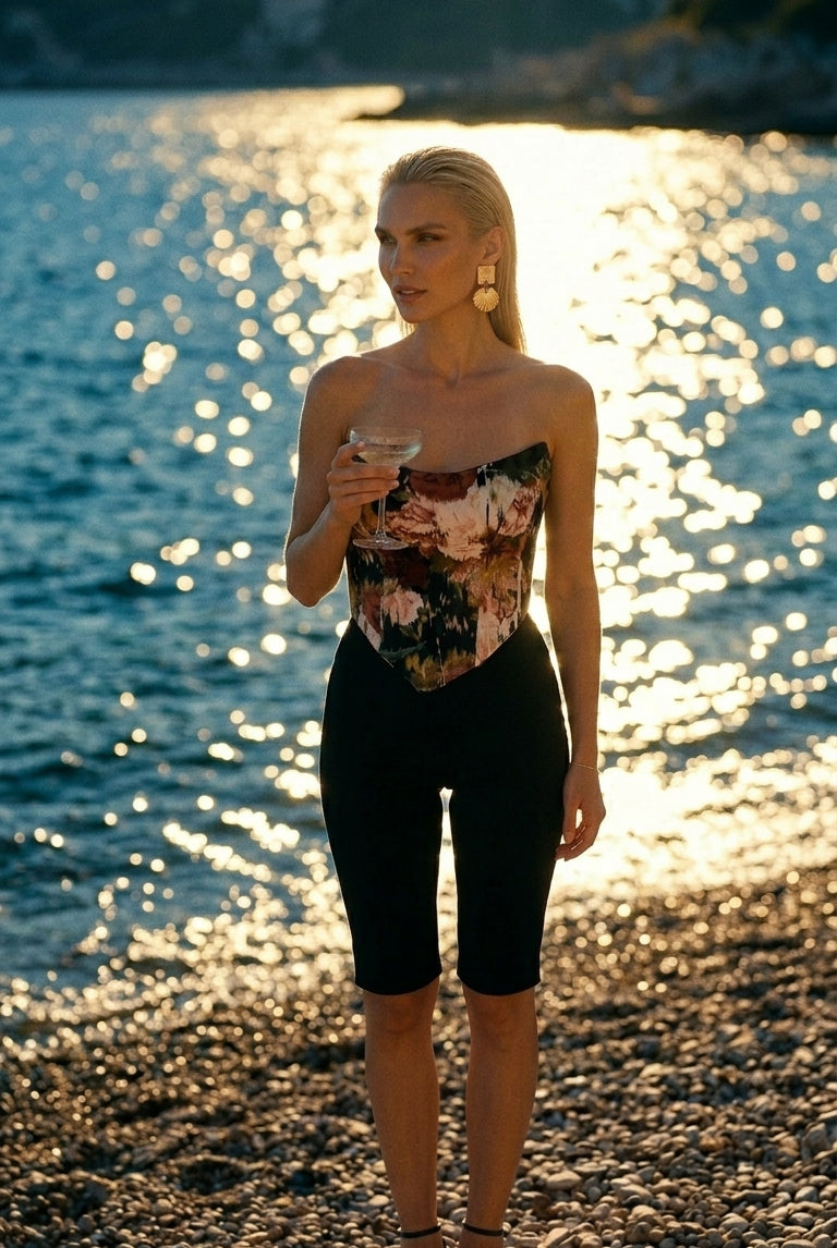 Wearing the Sarvin Ivy Corset Top, black shorts, and sandals, a woman stands on a rocky beach at sunset, holding a drink as sunlight glimmers on the water behind her.