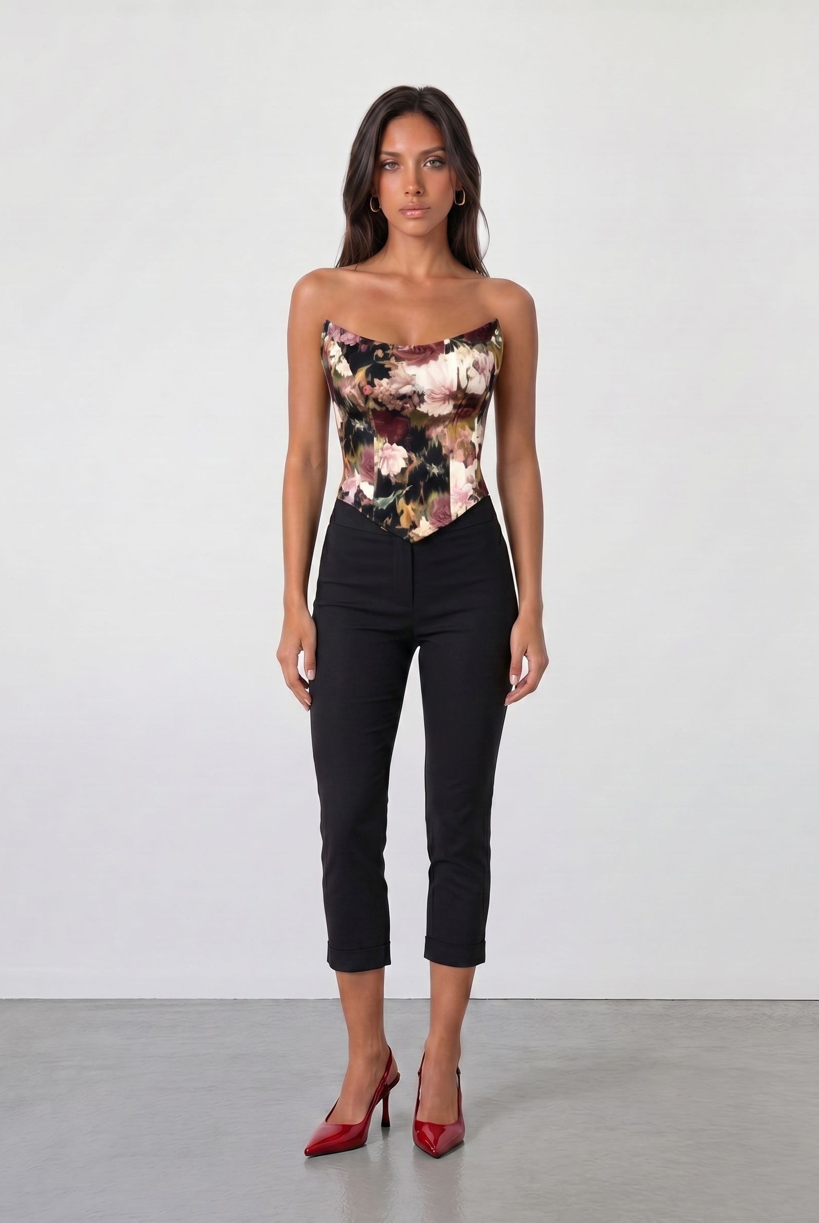A woman with long dark hair faces forward against a plain light wall, wearing the Sarvin Ivy Corset Top with a waist-cinching floral print, paired with black cropped pants and red high heels. Her expression is neutral.