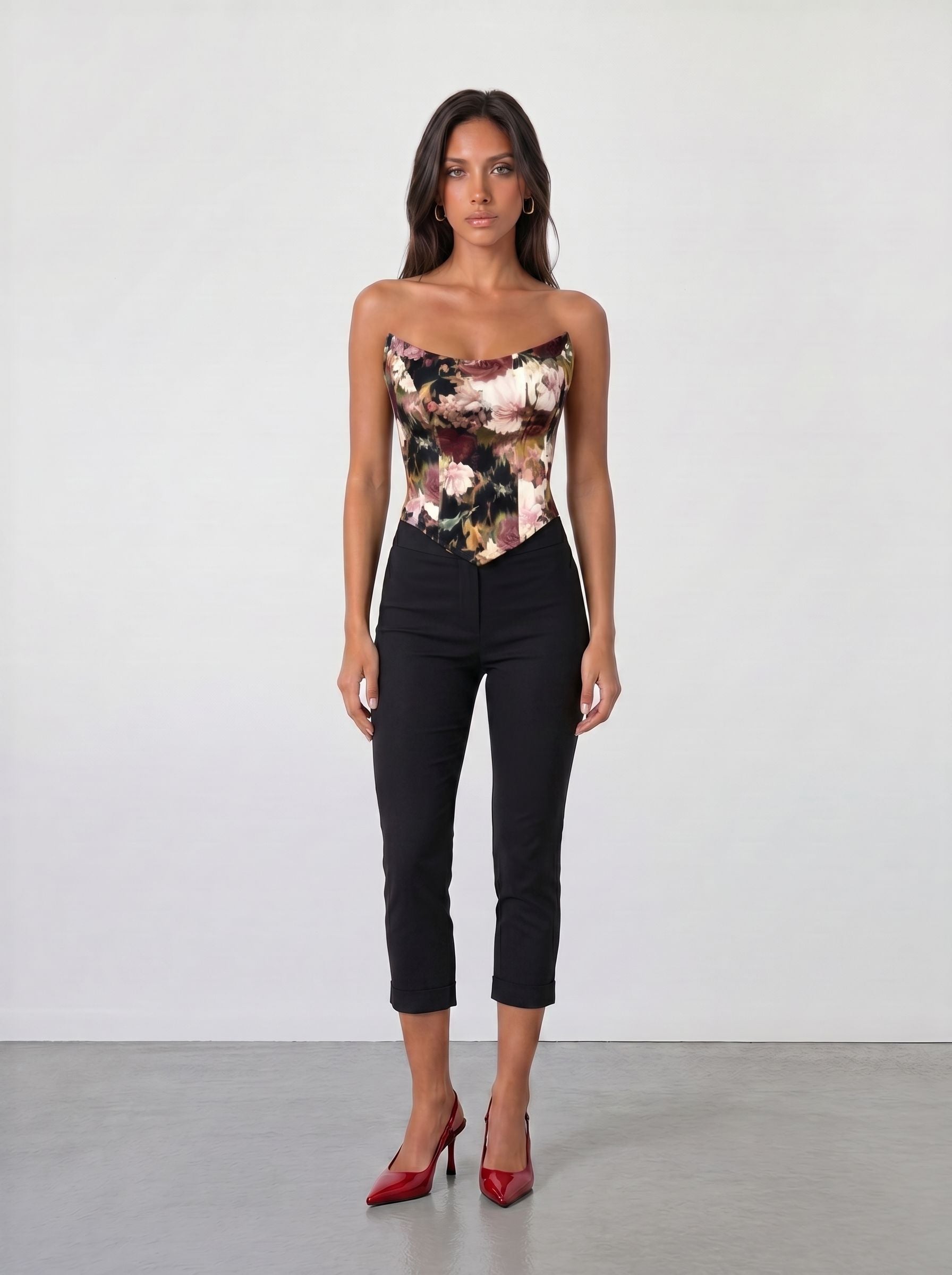 A woman with long dark hair faces forward against a plain light wall, wearing the Sarvin Ivy Corset Top with a waist-cinching floral print, paired with black cropped pants and red high heels. Her expression is neutral.