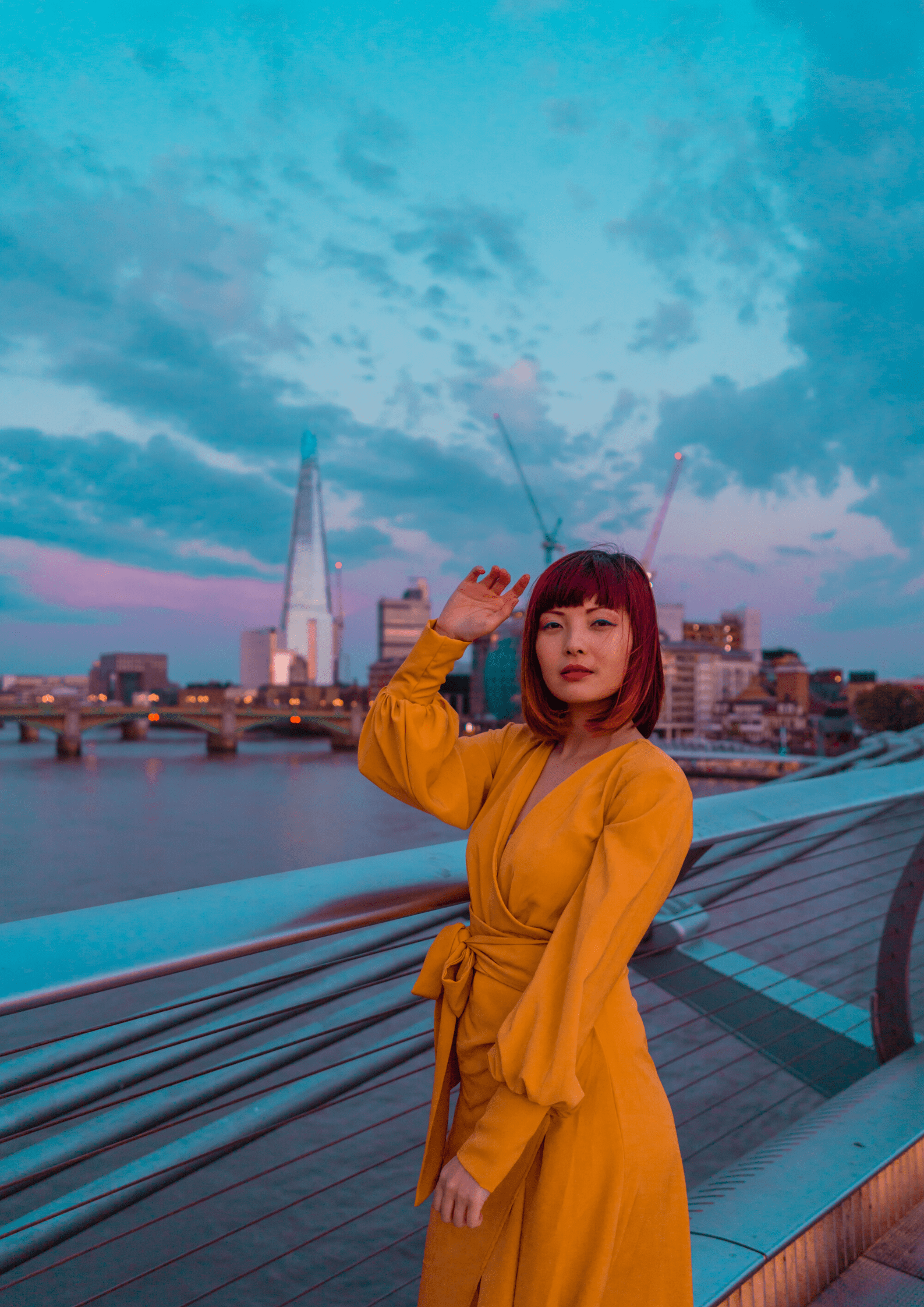 Wearing the Sarvin Mustard Wrap Maxi Dress with balloon bishop sleeves, a person stands on a bridge at sunset, city skyline and the Shard visible in the background under a cloudy sky.